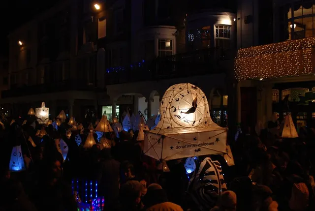 A giant illuminated clock-face lantern carried through a crowd of smaller lantern-bearers on a Brighton street at night during the Burning the Clocks parade.
