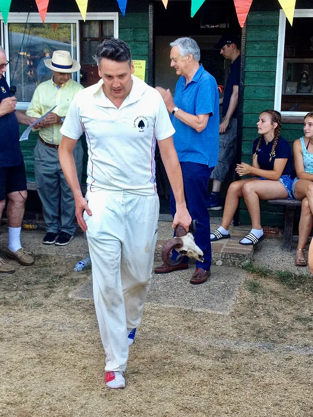 A cricketer in whites carries the pair of ram's horns awarded to the top scorer past bunting reading 'Horn Fair'.
