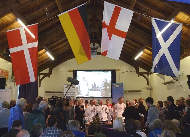 Cooks in whites are watched by a crowd beneath a row of national flags at the world porridge championships in Carrbridge village hall.