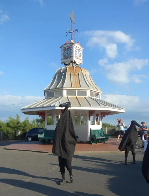 Two hooden horse performers in black robes stand beside the clock tower on Broadstairs seafront.