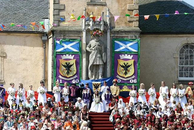 The Lanimer Queen and her court in ceremonial robes stand on the Mercat Cross steps above a crowd in Lanark.
