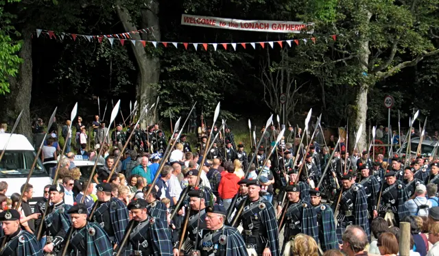 Rows of kilted Lonach Highlanders carrying raised pikes march past a 'Welcome to the Lonach Gathering' banner through a crowd.