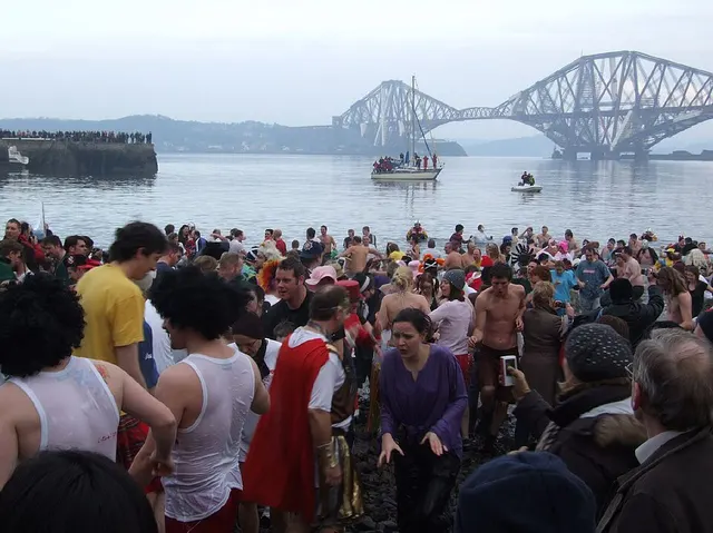 A large crowd in fancy dress wades into the Firth of Forth at South Queensferry on New Year's Day, with the Forth Bridge behind.
