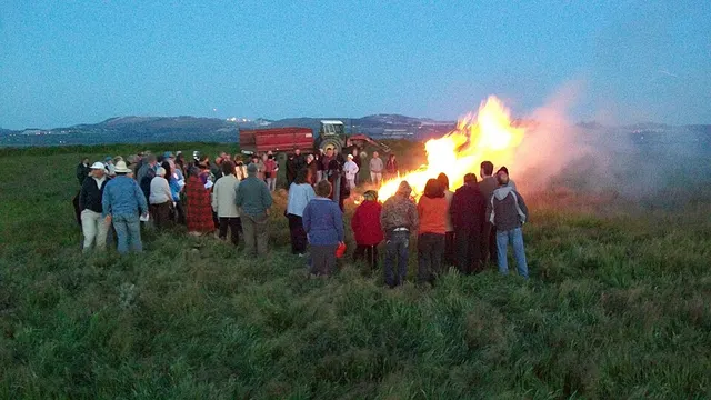 A crowd gathers around a blazing hilltop bonfire at dusk during Cornwall's midsummer bonfire chain.