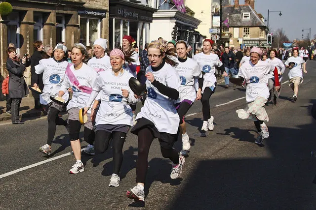 Women in aprons and headscarves sprint down Olney High Street carrying frying pans during the annual pancake race, watched by a crowd.