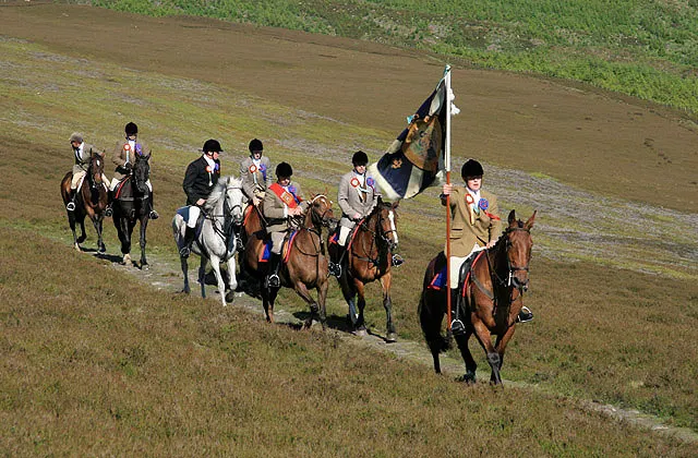 A standard bearer carrying a flag leads a line of mounted riders down a hillside track towards Selkirk during the Common Riding.