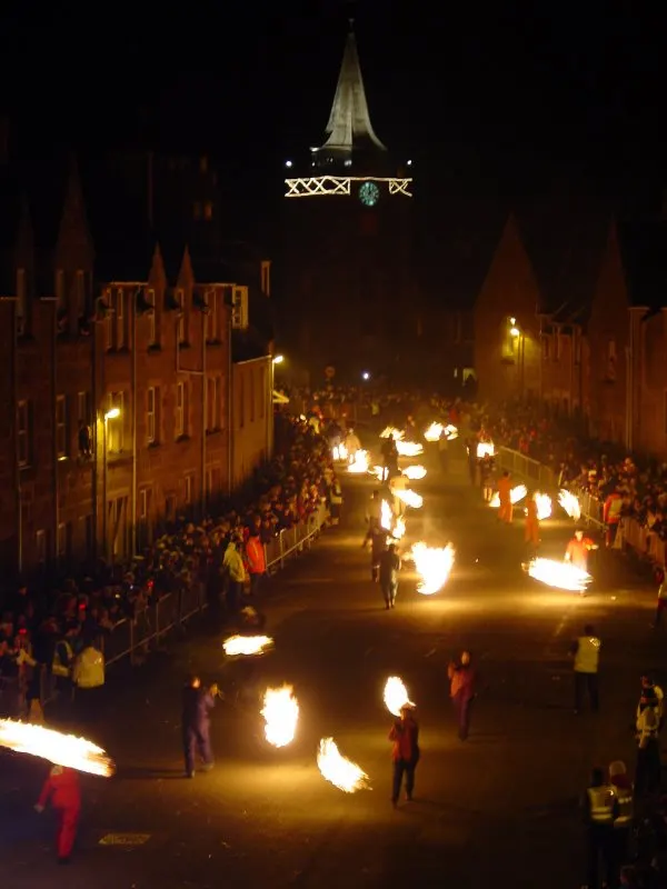Fire-swingers process up a crowded High Street in Stonehaven, swinging blazing balls of fire, with the Town House clock tower ahead.