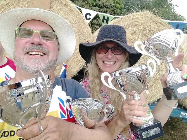 Two smiling competitors hold up trophies in front of hay bales and bunting at the World Pea Shooting Championships in Witcham.