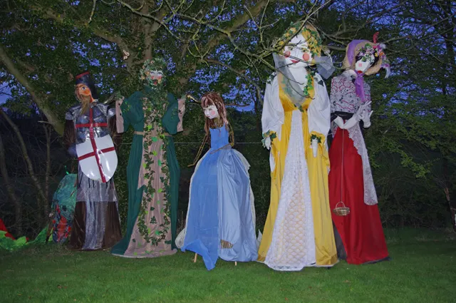 A group of costumed character scarecrows, including a knight and princesses, stands on display in a garden during the Wray Scarecrow Festival.