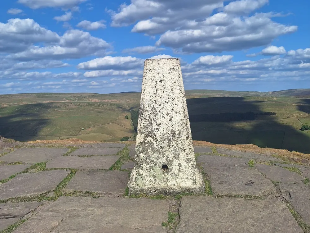 Photo of a white-painted Ordnance Survey trig pillar on a paved summit above moorland in the Peak District
